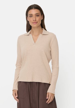 Kvinde med langt brunt hår iført en beige ribbet langærmet top med krave og brun nederdel mod en ensfarvet baggrund.