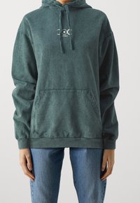 Mörkgrön hoodie med en framficka, dragsko i huvan och en liten vit logotyp på bröstet. Mjukt, texturerat tyg och avslappnad passform.