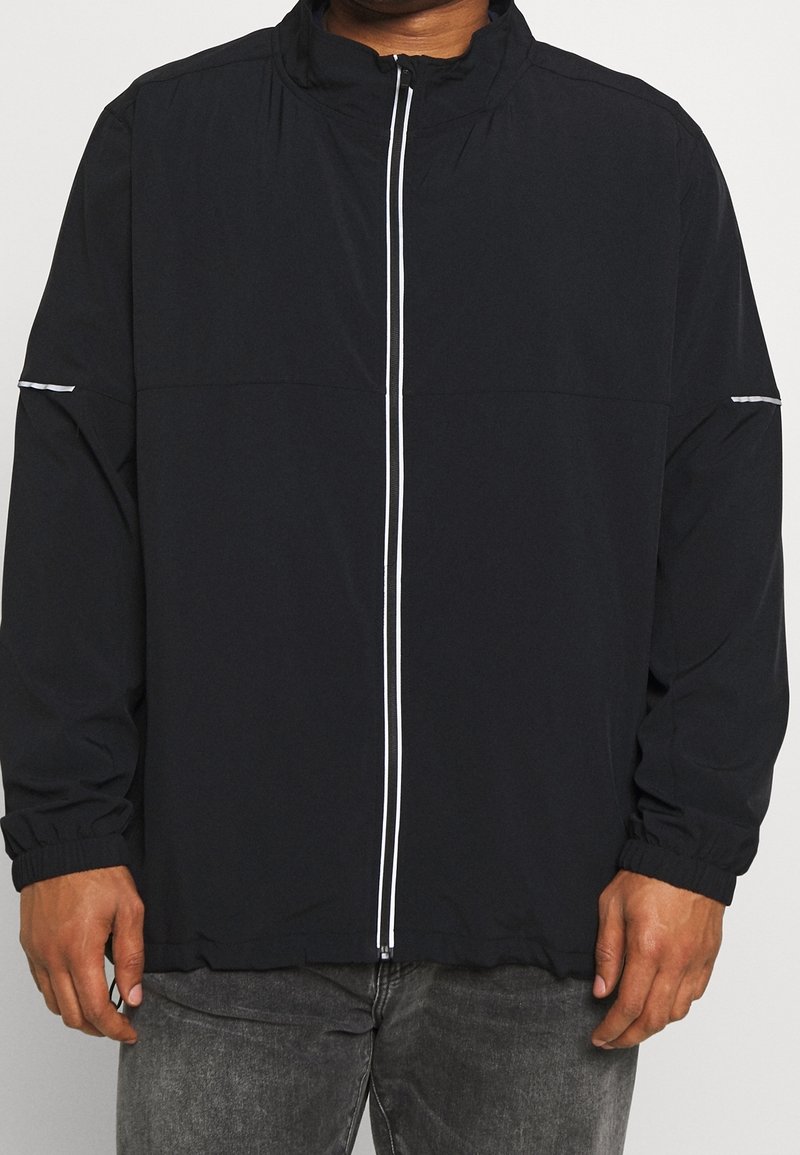 Homme portant une veste noire zippée avec une fermeture éclair blanche et des bandes réfléchissantes sur les manches, associée à un jean gris foncé.