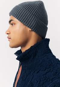 Siva rebrasta pletena kapa s fitiranom izradom. Nosi se s tamnoplavom teksturiranom flis jaknom s visokim ovratnikom, prikazano u profilu na svijetloj pozadini.