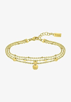 Pulsera en tono dorado que presenta cadenas superpuestas, charms esféricos y un cierre de gancho. Un detalle notable incluye una pequeña etiqueta grabada con "BOSS."