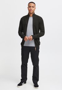 Cárdigan de punto verde oscuro con cuello alto, combinado con una camiseta gris clara. Pantalones negros y zapatillas negras con rayas blancas.