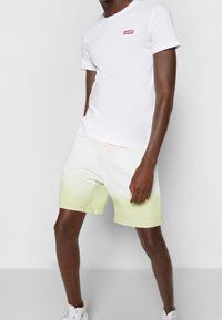 T-shirt blanc avec logo rouge, associé à un short dégradé de vert clair à blanc. Le short présente une texture lisse et une coupe décontractée.
