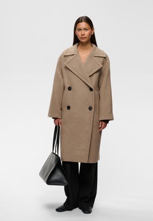 Femme portant un long manteau beige croisé, pantalon noir, chaussures noires, tenant un grand sac à main noir sur un fond clair uni.