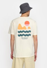 Camiseta de algodón crema con un gráfico de montañas y un sol en naranja y rosa, olas en azul oscuro, con el texto "Revolución" debajo.