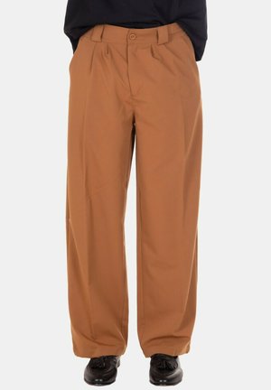 Pantaloni marrone cammello a gamba larga con pieghe anteriori, indossati con mocassini neri con nappine su sfondo bianco.