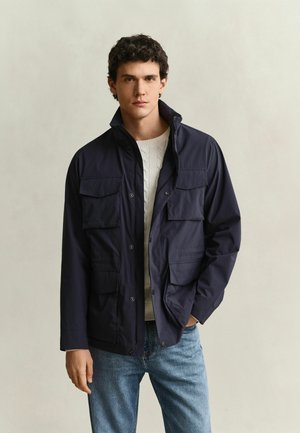 Man wearing a navy blue jacket with multiple front pockets over a white cable-knit sweater and blue jeans, standing against a plain background.