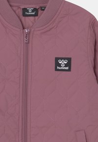 Kurtka bomberka pikowana w stonowanym mauve, z zamkiem błyskawicznym, ściągaczami na rękawach i czarną naszywką z logo na klatce piersiowej. Teksturalny wzór w romby.