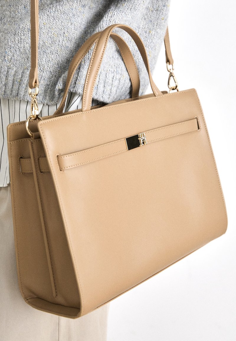 Sac à main en cuir structuré beige avec deux poignées et une bandoulière détachable, porté sur un pull et une chemise à rayures.