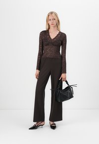 Brun blonde bluse med lange ermer og blomstermønster, kombinert med høyt liv mørke bukser. Modellen holder en liten svart veske med rynkete håndtak.