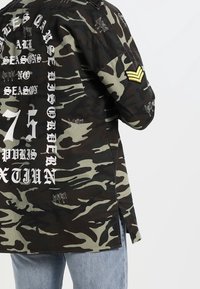 Giacca mimetica verde scuro e marrone con scritte bianche stampate, maniche lunghe, spacchi laterali e toppa gialla da sergente sul braccio sinistro.