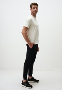 Herren T-Shirt mit kurzen Ärmeln in Creme, kombiniert mit schwarzen tapered Pants mit Taschen. Das Model trägt schwarze Sneaker mit strukturierten Akzenten.