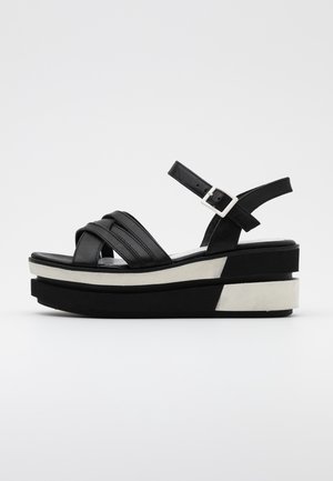 Sort læderplatformsandal med hvide såleaccents, justerbar ankelrem og krydsede frontstropper på en neutral baggrund.