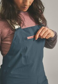 Blue overall with a textured finish features a chest pocket with a pink zipper. A long-sleeve pink shirt is worn underneath.