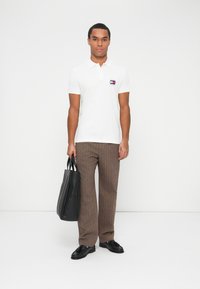 Uomo con camicia polo bianca, pantaloni a righe marroni, mocassini neri, che tiene una borsa a tracolla in pelle nera, in piedi contro uno sfondo bianco.