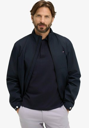 Homme barbu portant une veste bleu marine sur un polo foncé et un pantalon gris clair, debout les mains dans les poches devant un fond blanc.