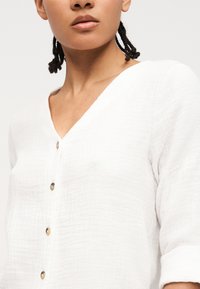 Gros plan d'une personne vêtue d'un haut blanc texturé à boutons avec des manches retroussées et des cheveux tressés, sur un fond uni.