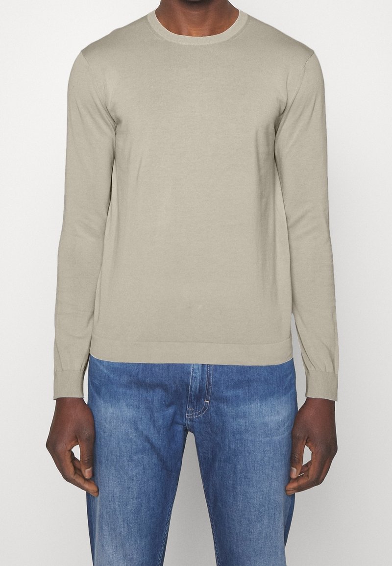 Hellbeige, langärmliger Pullover aus weichem Material mit rundem Halsausschnitt und gerippten Manschetten, kombiniert mit blauen Jeans.