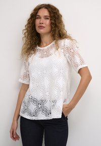 T-shirt bianco in pizzo con motivi floreali, maniche corte e vestibilità ampia. Il materiale appare leggero con una finitura testurizzata e semi-trasparente.
