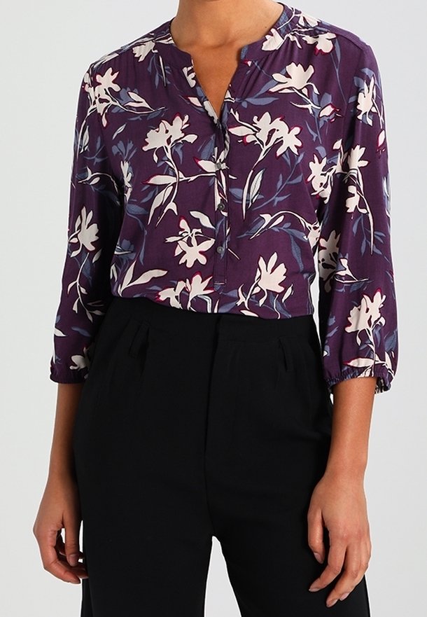 Blouse violette avec imprimé floral blanc et rose, dotée d'un col boutonné et de manches trois-quarts, associée à un pantalon noir.