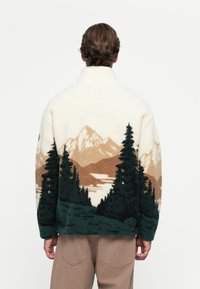 Man wearing a fleece jacket with a mountain and forest landscape design on the back, standing against a plain white background.