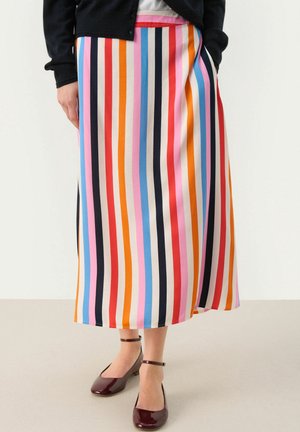 Kobieta w kolorowej, pionowo paskowanej spódnicy midi z ręką w kieszeni, czarnym kardiganie i bordowych butach na kostkę stoi na beżowej podłodze.