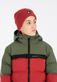 Giacca a vento imbottita rossa e verde con una fascia nera e logo, con chiusura a zip e cappuccio. Indossata con un berretto a coste rosso.