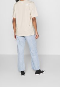 T-shirt beige a maniche corte con scollo rotondo, caratterizzato da un piccolo logo bianco sulla parte superiore della schiena, abbinato a jeans a gamba larga blu chiaro e sneaker nere.