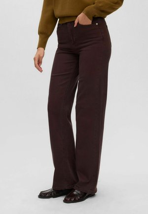 Persona che indossa pantaloni ampi marrone scuro, mocassini neri e un maglione verde oliva con una mano in tasca.
