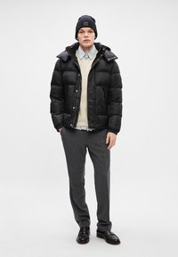 Μαύρο μπουφάν πούφ με γυαλιστερή υφή, ελαφρύ πουλόβερ και πουκάμισο με γιακά. Συνδυάζεται με γκρίζες παντελόνες και μαύρες δερμάτινες παντόφλες.