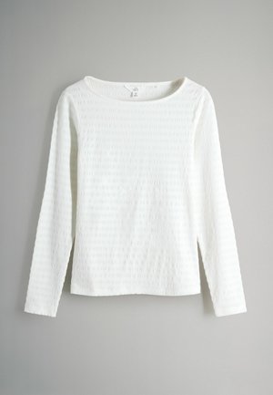Fehér hosszú ujjú felső texturált anyagból, széles nyakkivágással és egyenesen szabott fazonnal. Az anyag finom mintázott dizájnt mutat.