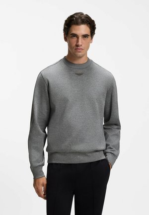 Mand iført en ensfarvet grå crewneck sweatshirt og sorte bukser, stående med den ene hånd i lommen mod en hvid baggrund.