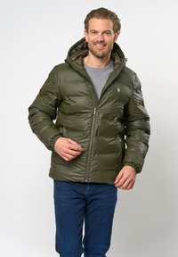 Žalia oliivinė dangaus striukė su kapišonu, su užtrauktuku, elastinėmis rankovėmis ir pilku pamušalu. Modelis vilki tamsiai mėlynas džinsus.