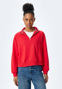 Czerwona bluza na zamek z stójką, z ribbowymi mankietami i dołem. Posiada biało-niebieski pasek na kołnierzu. Casualowy krój.