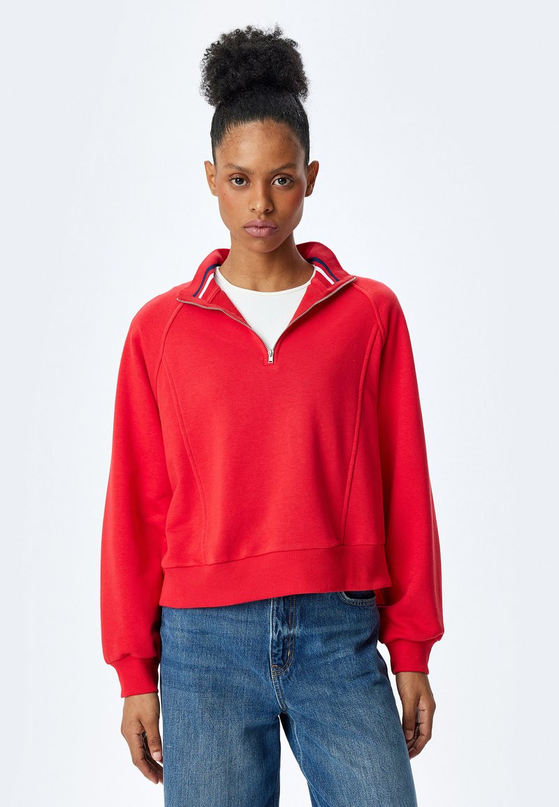 Czerwona bluza na zamek z stójką, z ribbowymi mankietami i dołem. Posiada biało-niebieski pasek na kołnierzu. Casualowy krój.