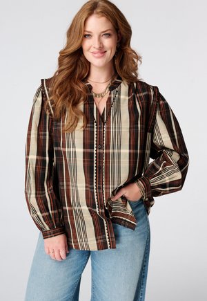Vrouw met lang golvend rood haar, gekleed in een bruin en beige geruit blouse met knopen en blauwe jeans, glimlachend met één hand in haar zak.
