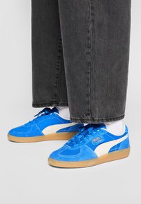 Baskets bleues en daim et tissu avec des accents blancs et une semelle en caoutchouc, associées à un jean large gris et des chaussettes blanches.