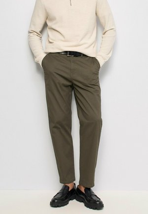 Hombre con camiseta beige de manga larga, pantalones rectos verde oliva con cinturón negro y zapatos de cuero negros gruesos, de pie con las manos en los bolsillos.