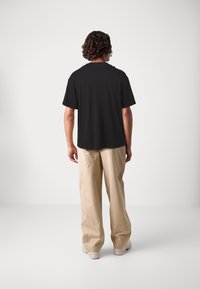 Zwarte T-shirt met korte mouwen en een relaxte pasvorm, gedragen met lichtbeige wijde broek en witte sneakers, gezien vanuit de achterkant.