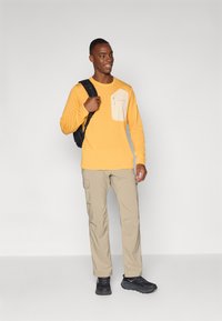 Langærmet gul skjorte med en beige lomme, parret med lyse beige cargo-bukser og sorte sneakers. Modellen bærer en sort rygsæk.