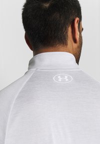 Graues Sportshirt mit hohem Kragen und glatter Textur, mit einem weißen Logo auf der Rückseite. Nahaufnahme von hinten.
