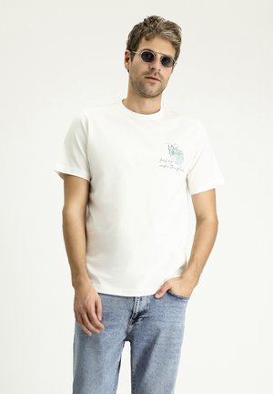 Camiseta de algodón blanca de manga corta, con un diseño gráfico verde en el pecho. Combina con unos vaqueros de mezclilla azul claro y gafas de sol redondas.