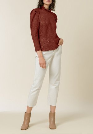 Kobieta stojąca w czerwonej koronkowej bluzce z długim rękawem, białych skróconych dżinsach i beżowych botkach, na tle jednolitego beżowego tła.