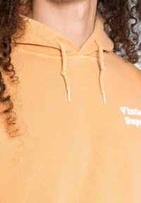 Hooded sweatshirt in light orange cotton. Features white embroidered text "Vintage" on the front and textured drawstring cords.