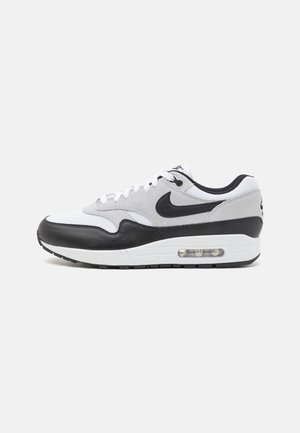 Basket Nike Air Max 1 avec une tige blanche et grise, des accents en cuir noir, une forme arrondie et un amorti visible dans la semelle.