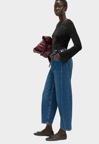 Czarna, teksturowana bluzka z długim rękawem, niebieskie spodnie typu wide-leg, czarne płaskie buty. Trzyma bordo torbę; wyróżnia się srebrnym okuciem na pasku.