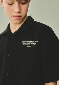 Polo negro con un diseño abotonado. En el pecho delantero tiene texto blanco que dice "TOKYO DISTRICT.JPN" junto con coordenadas y caracteres japoneses.