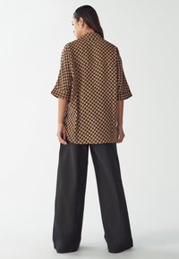 Bruine blouse met een geometrisch patroon, korte mouwen en een losse pasvorm, gecombineerd met zwarte wijde pantalons. Zachte stofstructuur.