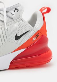 Szürke sportcipő hálós felsőrésszel, fekete Nike logóval, narancssárga részletekkel és egy feltűnő piros, légpárnás talprészel.