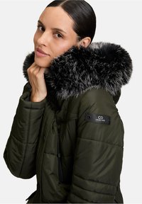 Oliwkowa kurtka puchowa z kapturem z czarno-szarego sztucznego futra, z zamkiem błyskawicznym w kieszeni oraz naszywką z logo na rękawie. Gładka tekstura.